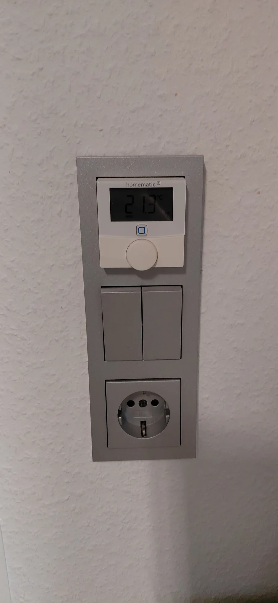 Homematic IP Wandthermostat in einem Schalterrahmen über zwei Lichtschaltern und einer Steckdose, Display zeigt 21,3 °C